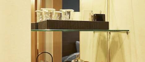 A kitchenette with a coffee maker, kettle, and a glass shelf with cups and a container of Pringles.