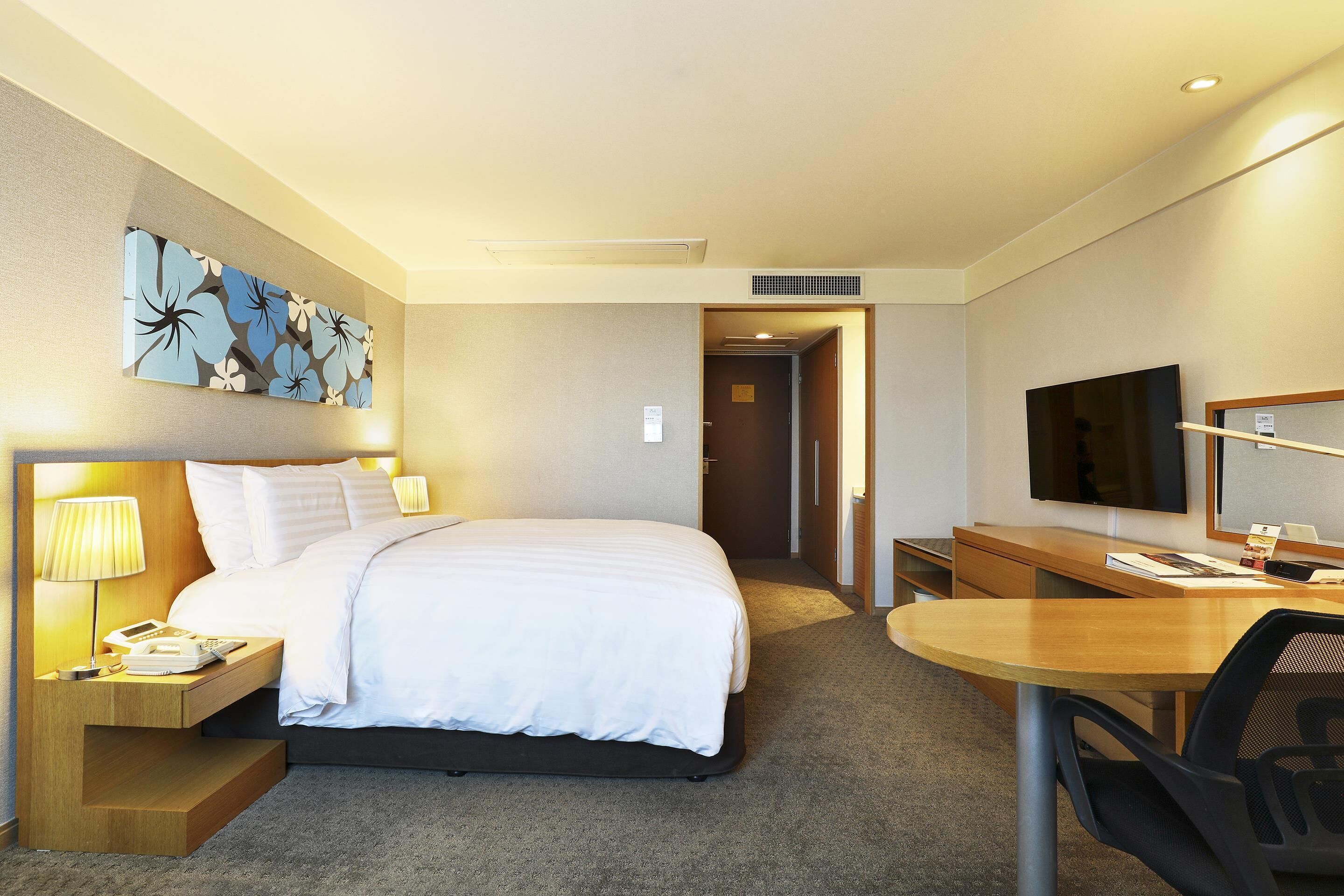 A hotel room with a bed, bedside table, lamp, desk, chair, television, and a painting on the wall.
