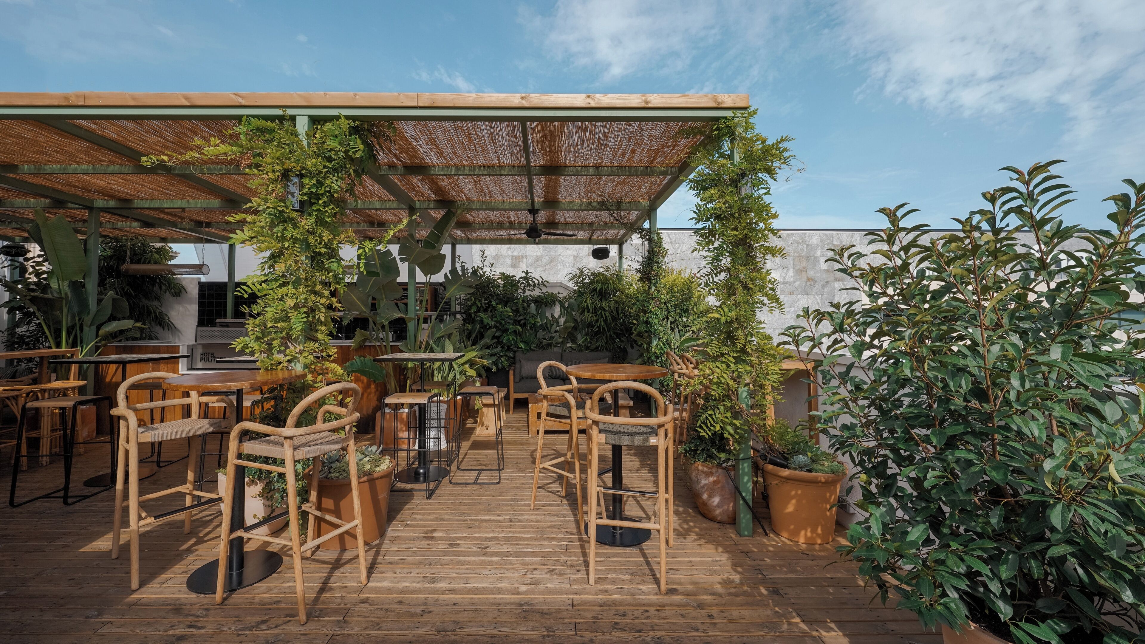 An outdoor seating area with wooden tables and chairs, surrounded by potted plants and a pergola structure.
