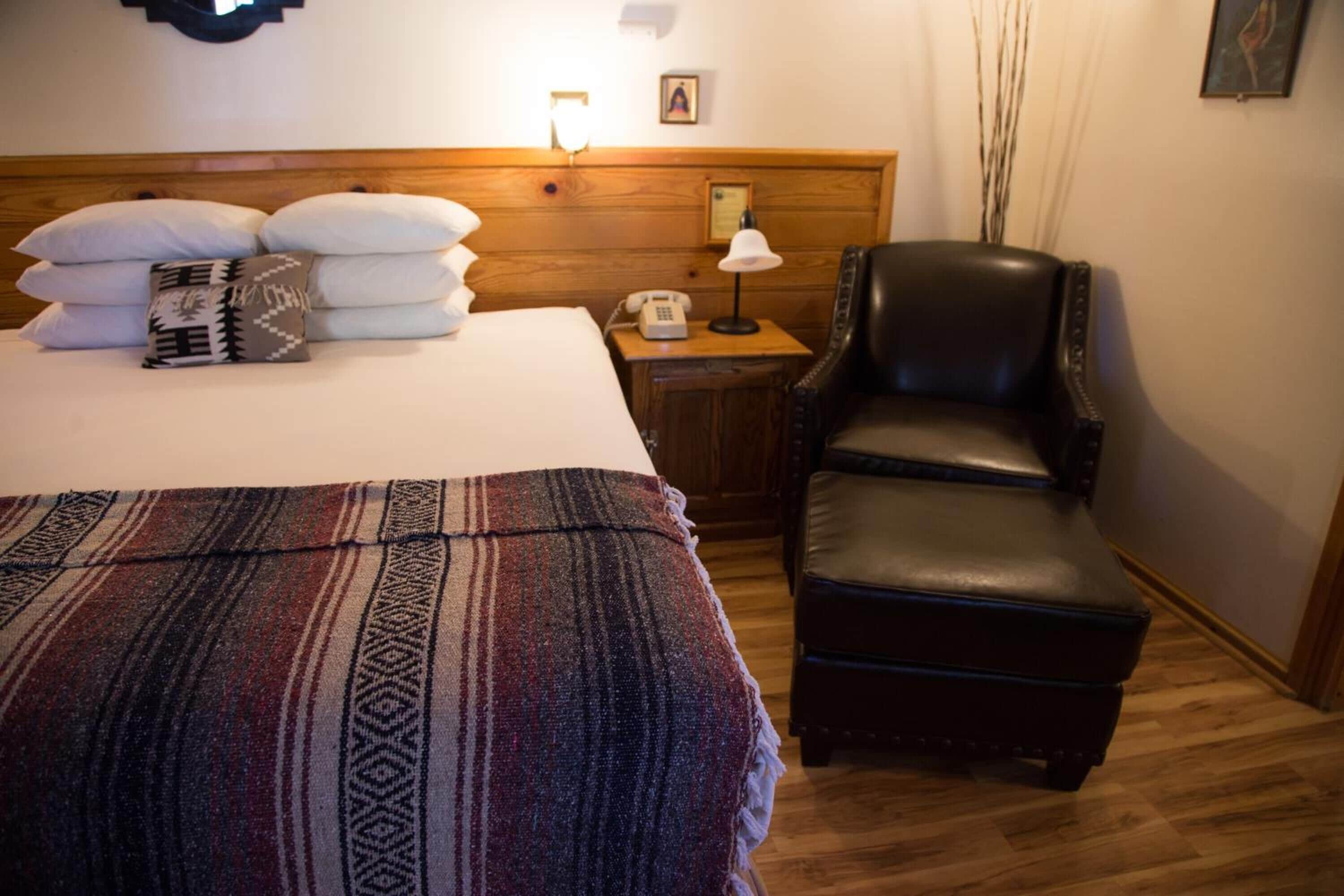 Habitación de hotel con cama, una silla, mesita de noche con lámpara y cabecero de madera.