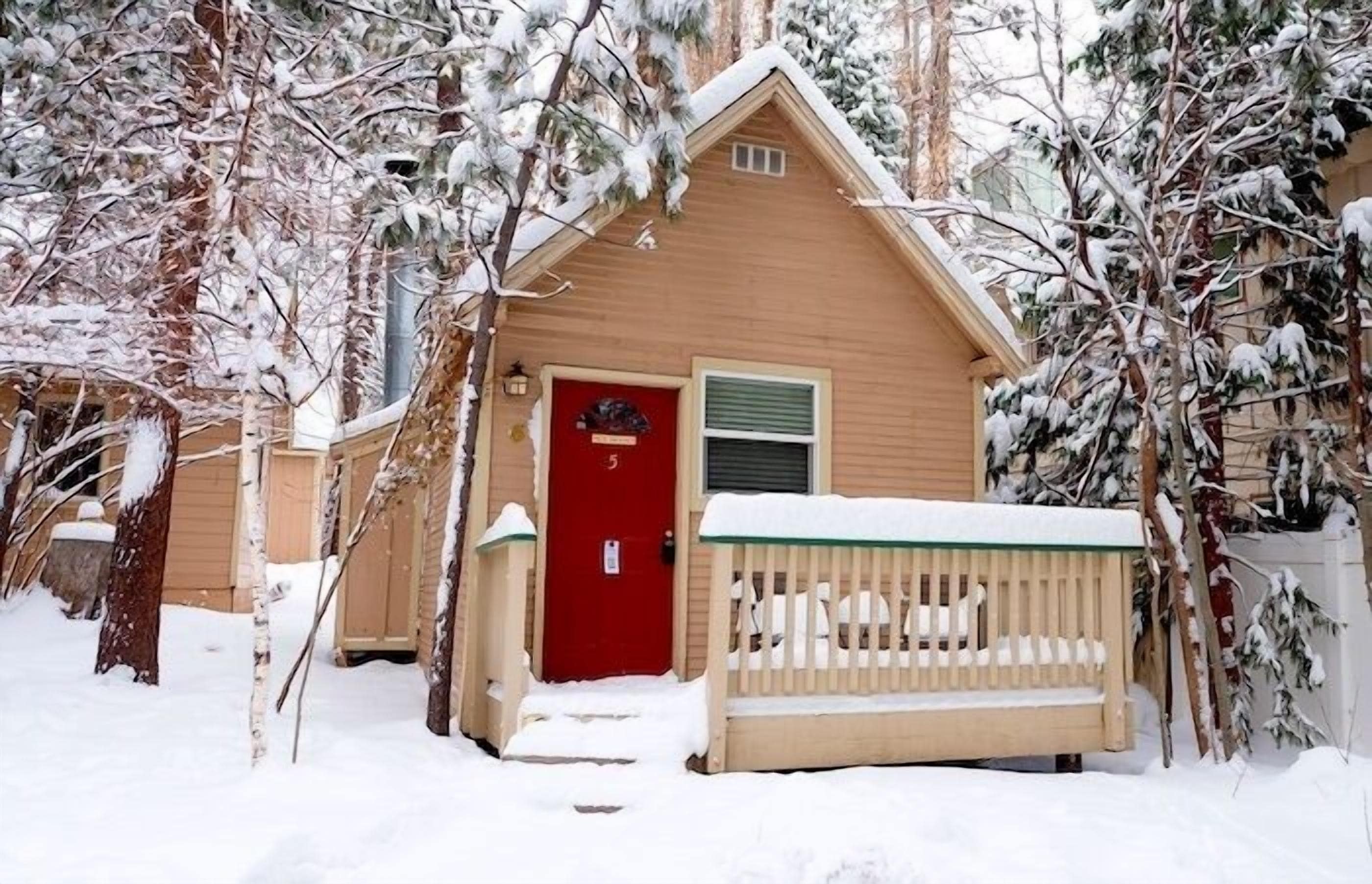 Ein kleines, eingeschossiges Haus mit roter Tür, umgeben von schneebedeckten Bäumen.