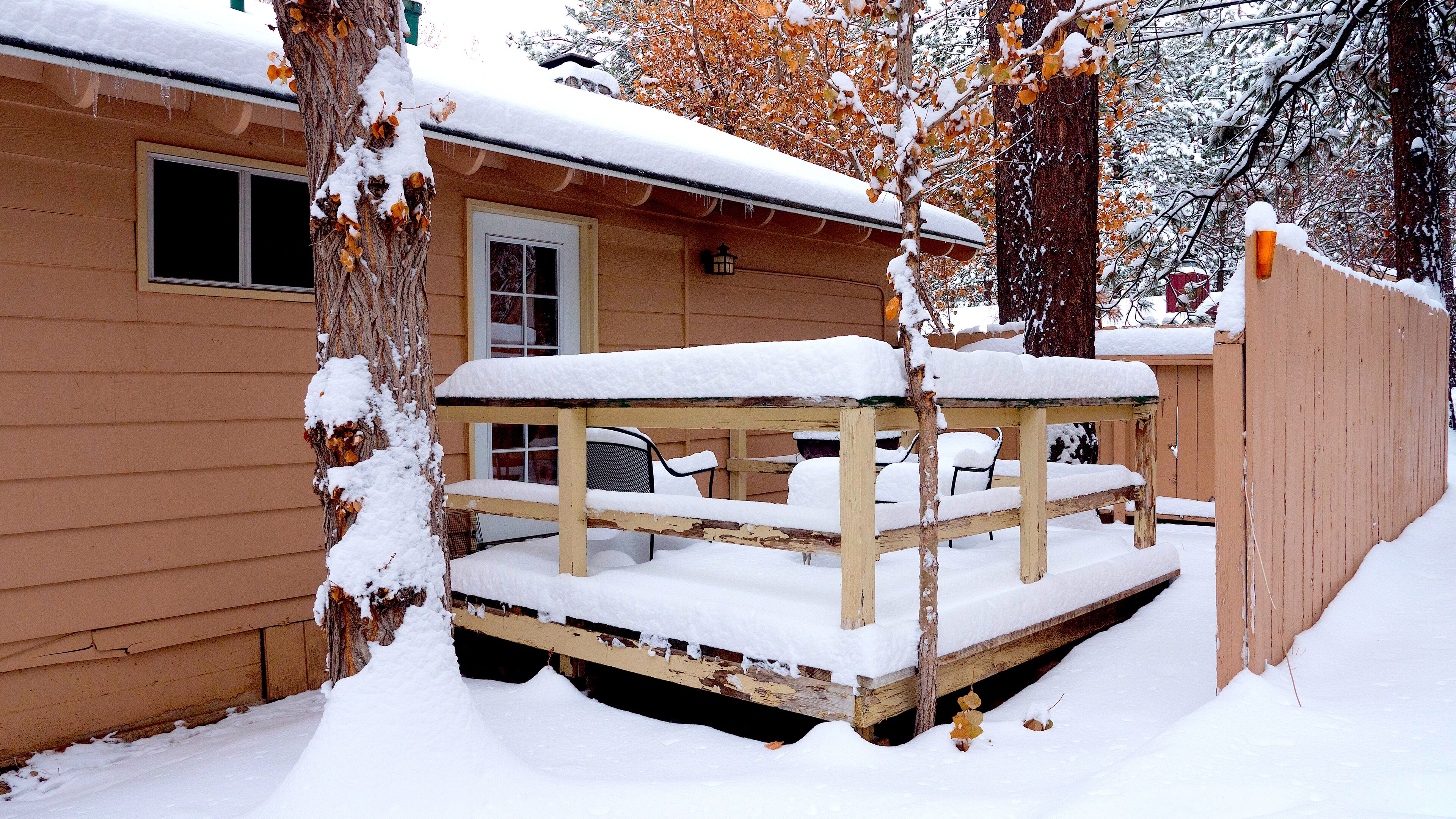 A wooden deck with a bench and a table covered in snow, adjacent to a house with a door and windows.