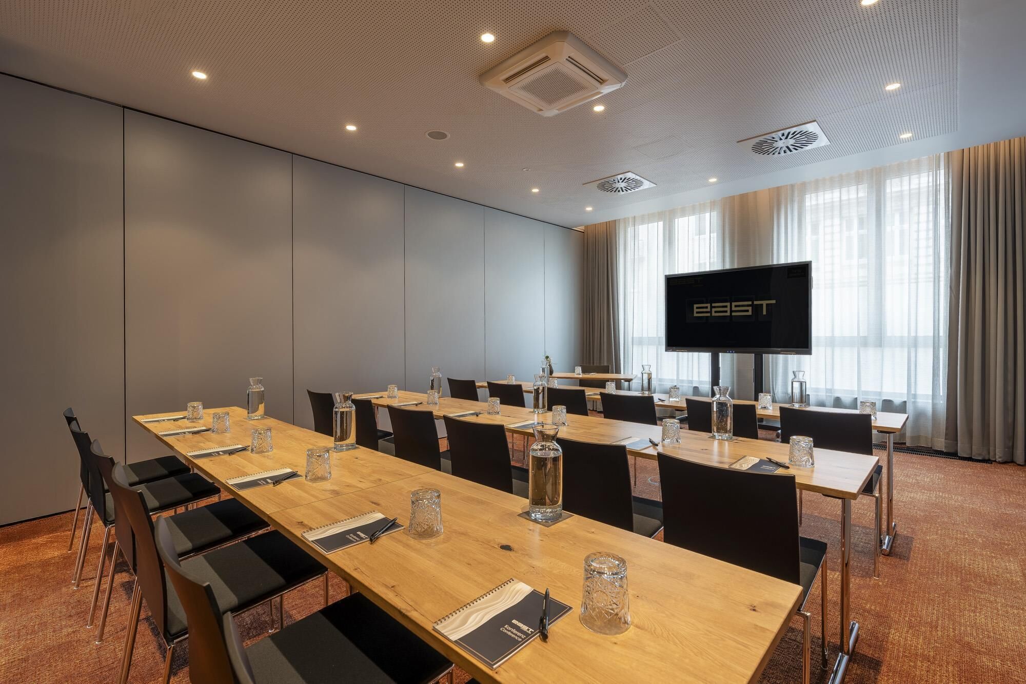 Una sala de conferencias con una mesa larga, sillas, un televisor de pantalla plana y un ventanal con cortinas.