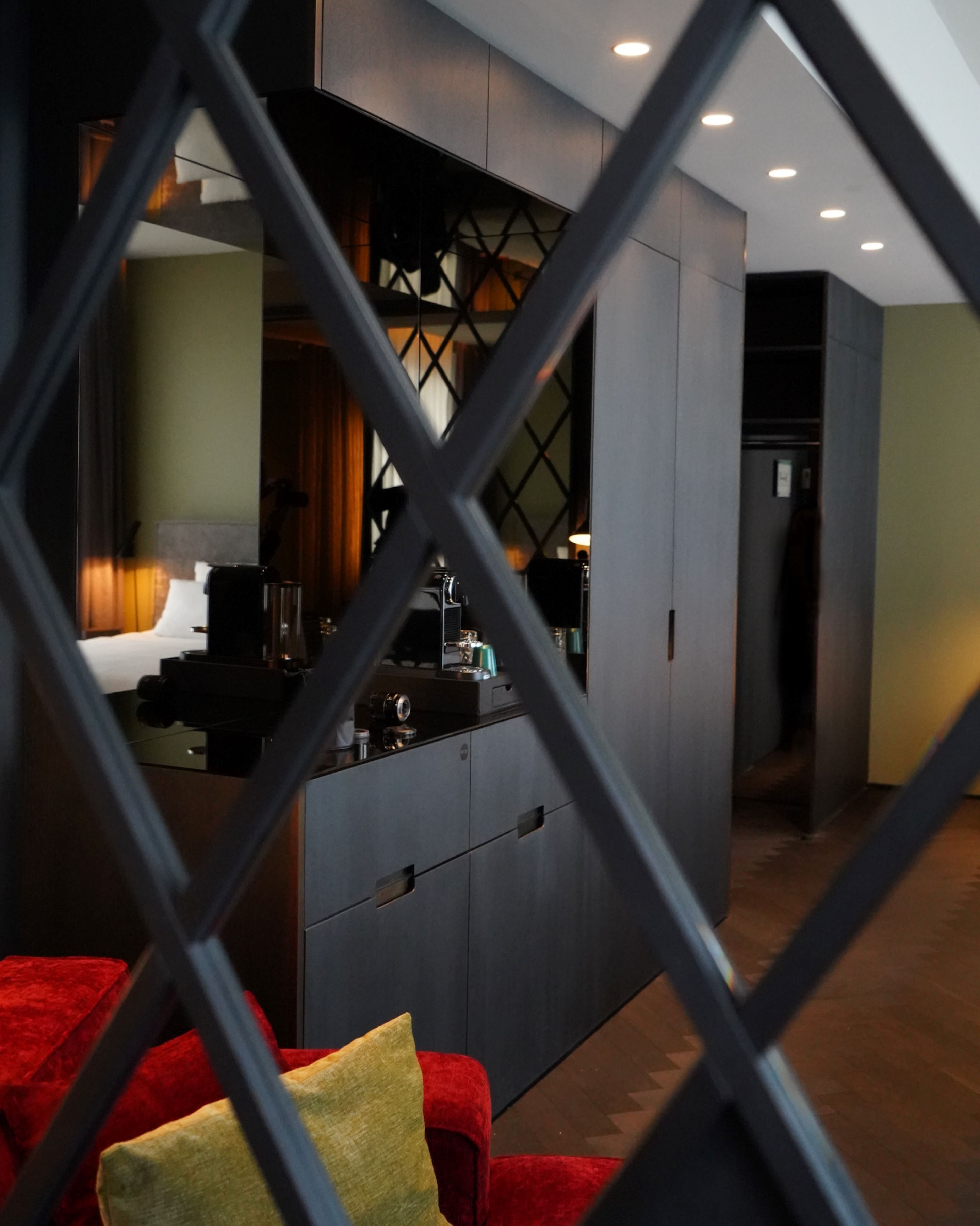 Ein modernes Hotelzimmer mit einem dunkelbraunen Kleiderschrank, einem roten Sofa mit einem gelben Kissen und einem durch eine geometrisch gemusterten Trennwand erkennbaren Küchenbereich.