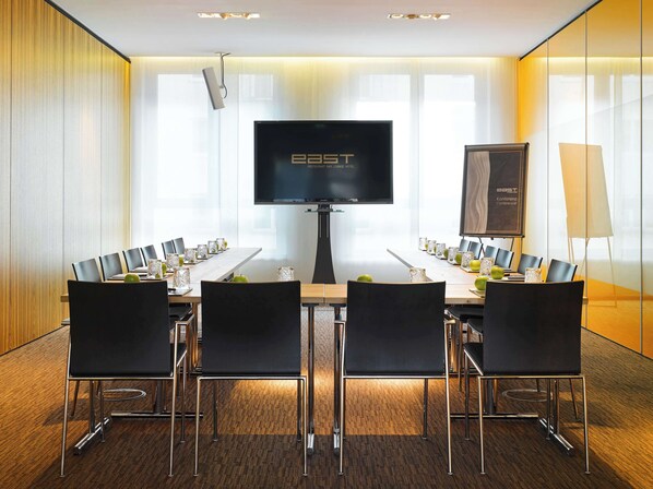 Un salón de conferencias con una gran televisión de pantalla plana, un proyector y una pantalla para presentaciones. Hay varias sillas alrededor de una mesa con vasos y manzanas.