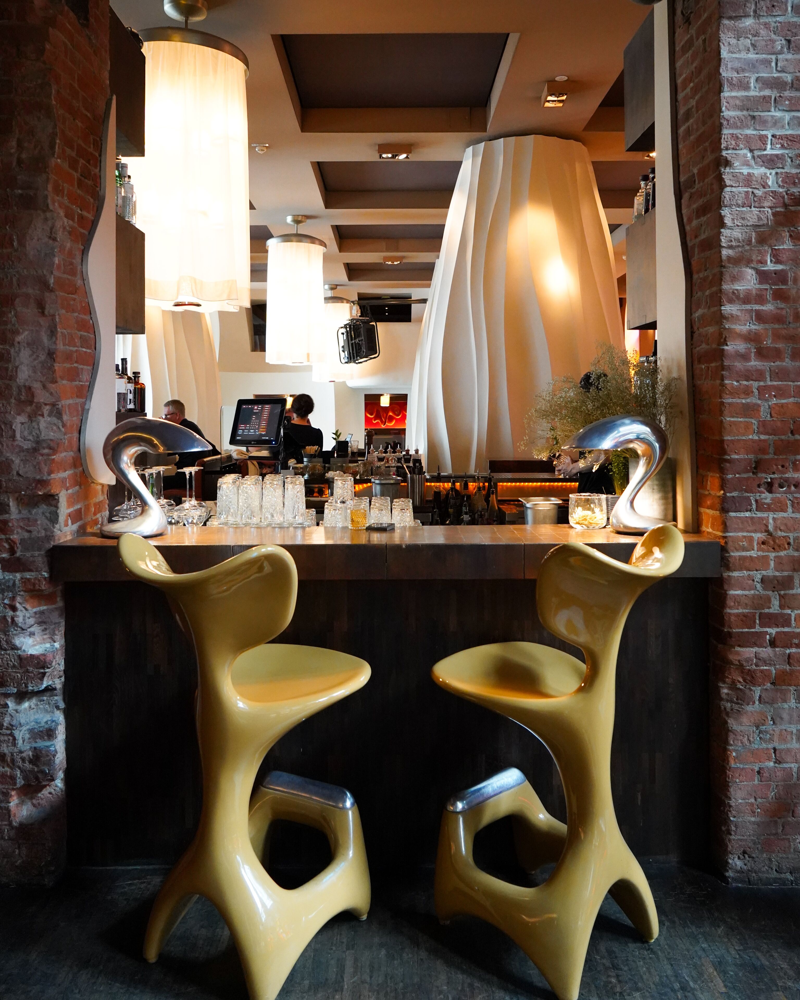 A bar area with two yellow chairs, a counter with a digital screen, and a brick wall.