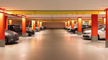 Eine mehrgeschossige Parkgarage mit in Reihen geparkten Autos, beleuchtet durch Deckenleuchten, und gestützt von orangefarbenen Säulen.