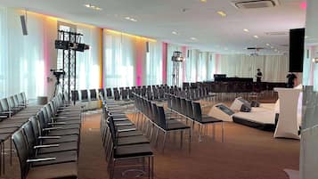 Ein Konferenzraum mit Reihen von Stühlen, einer Bühne mit Rednerpult und einem großen Fenster mit Vorhängen.