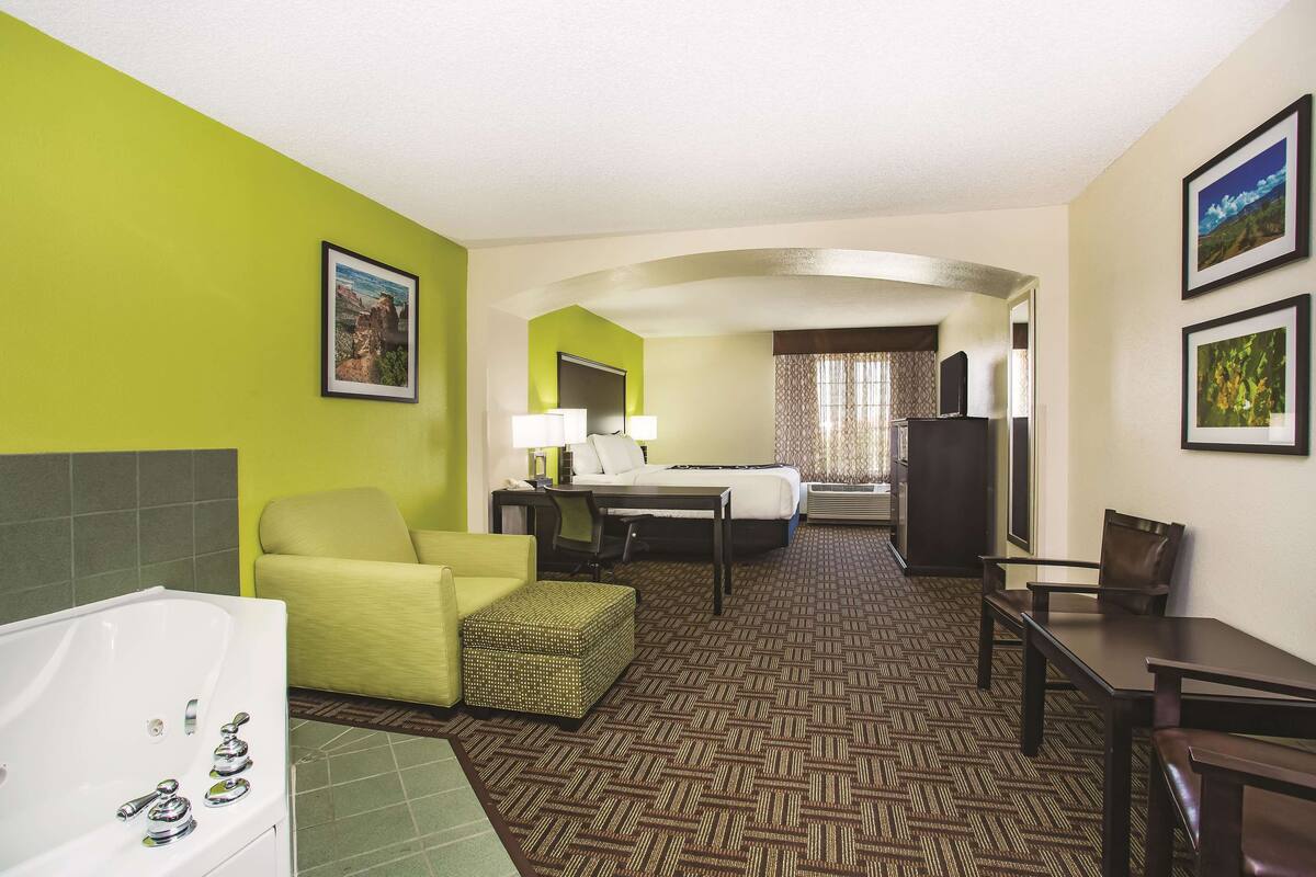 Ein Hotelzimmer mit grüner Wand, einem Bett, einem Schreibtisch, einem Stuhl, einem kleinen Tisch, einer Lampe, einem Spiegel und einer Badewanne.