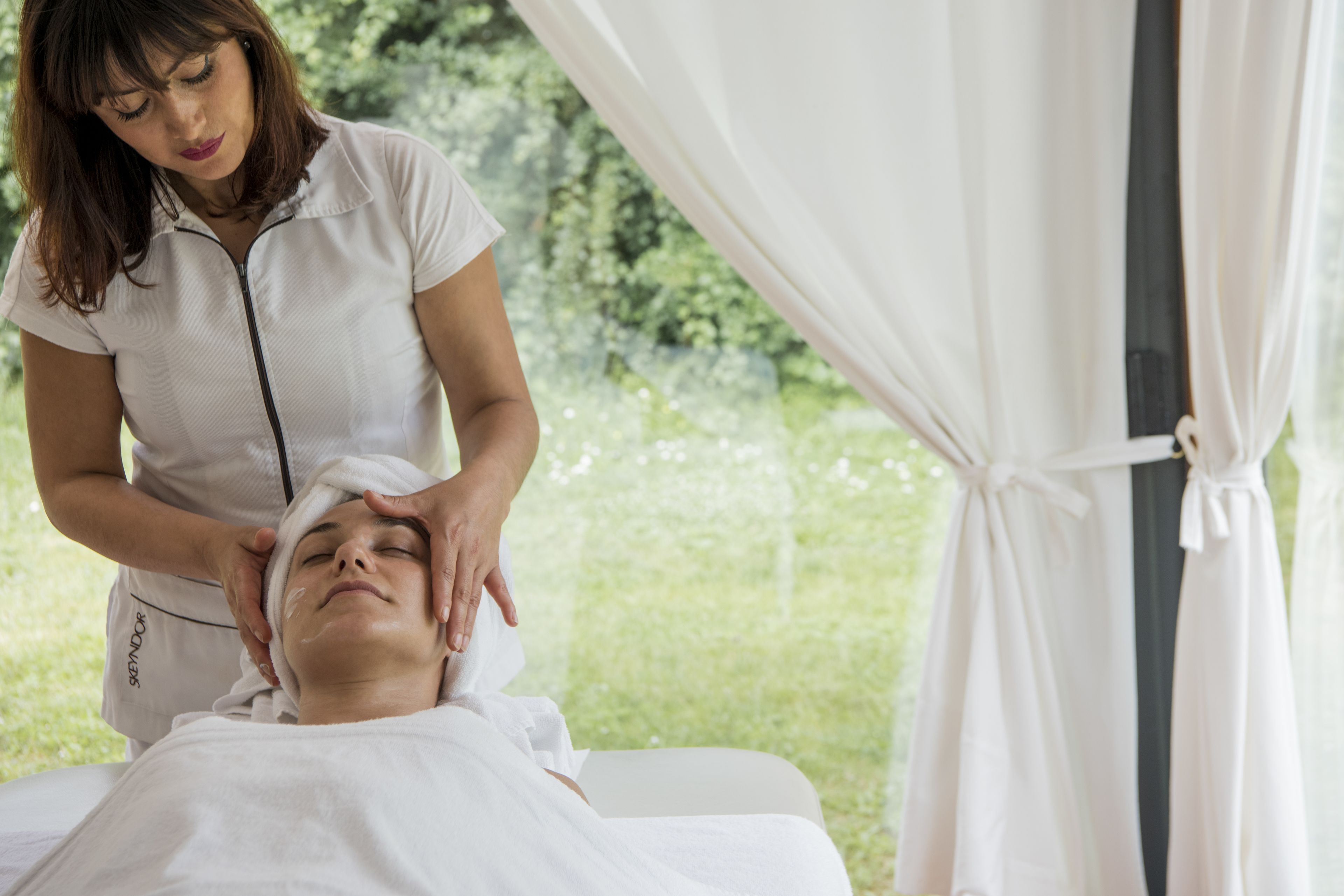 Une personne reçoit un massage de la tête dans une pièce d’où l’on peut admirer la verdure environnante.