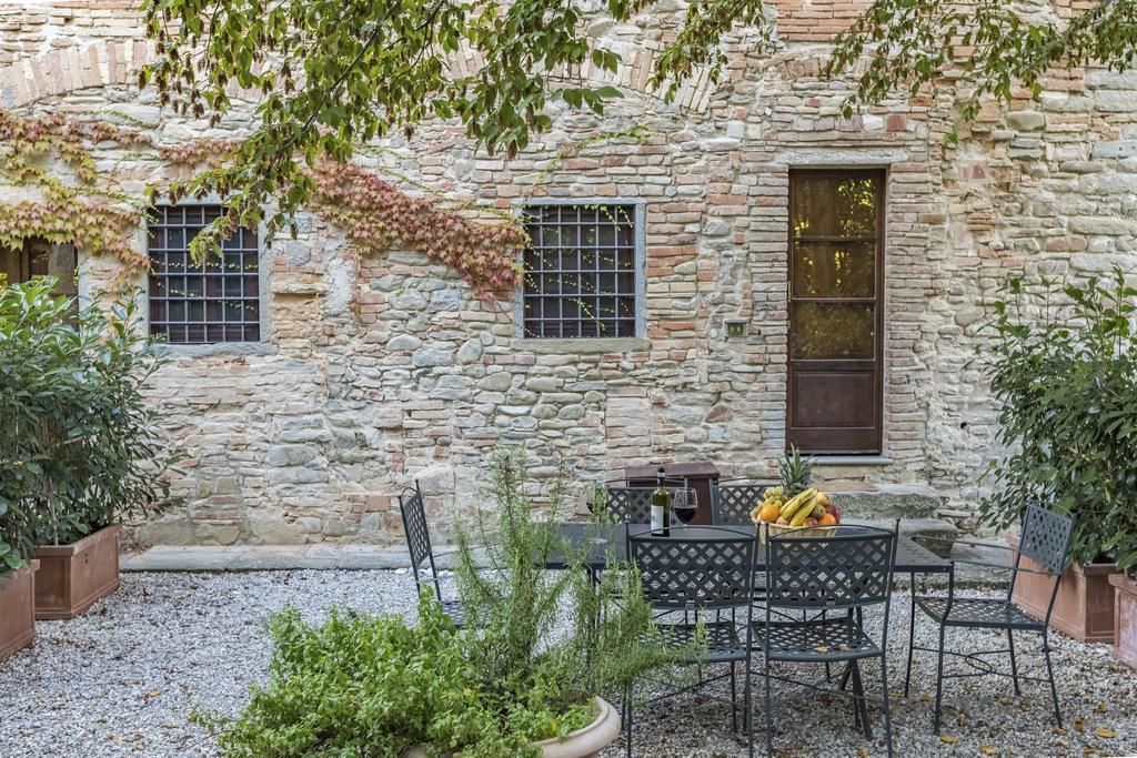 A stone building with a rustic exterior, featuring a small outdoor seating area with metal chairs and a table, surrounded by potted plants and a gravel ground.