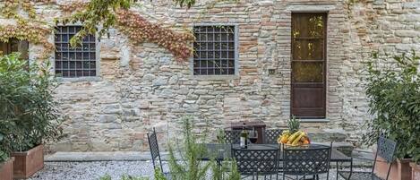 A stone building with a rustic exterior, featuring a small outdoor seating area with metal chairs and a table, surrounded by potted plants and a gravel ground.