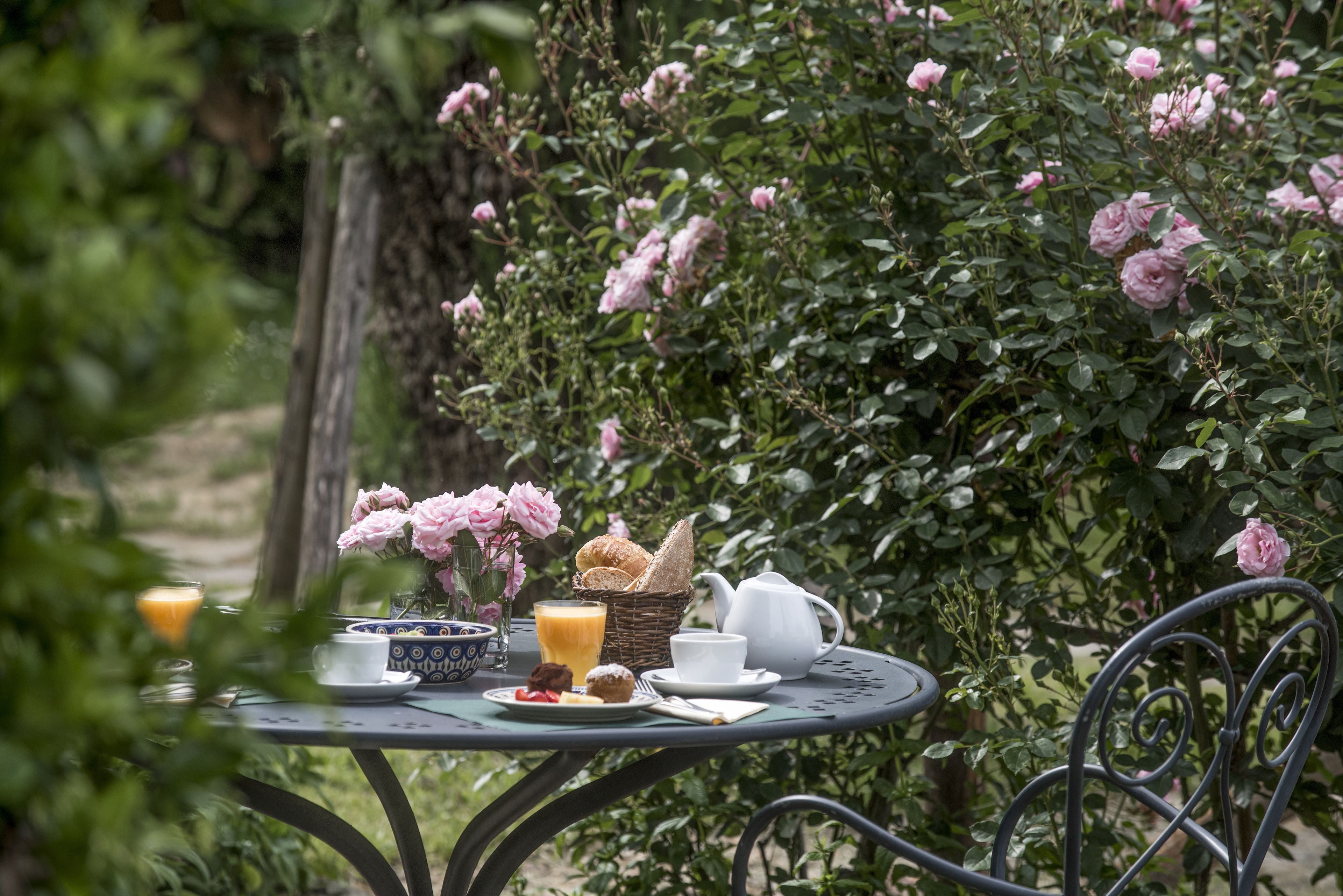 A table set with breakfast items including croissants, pastries, and a teapot, surrounded by lush greenery and pink roses.