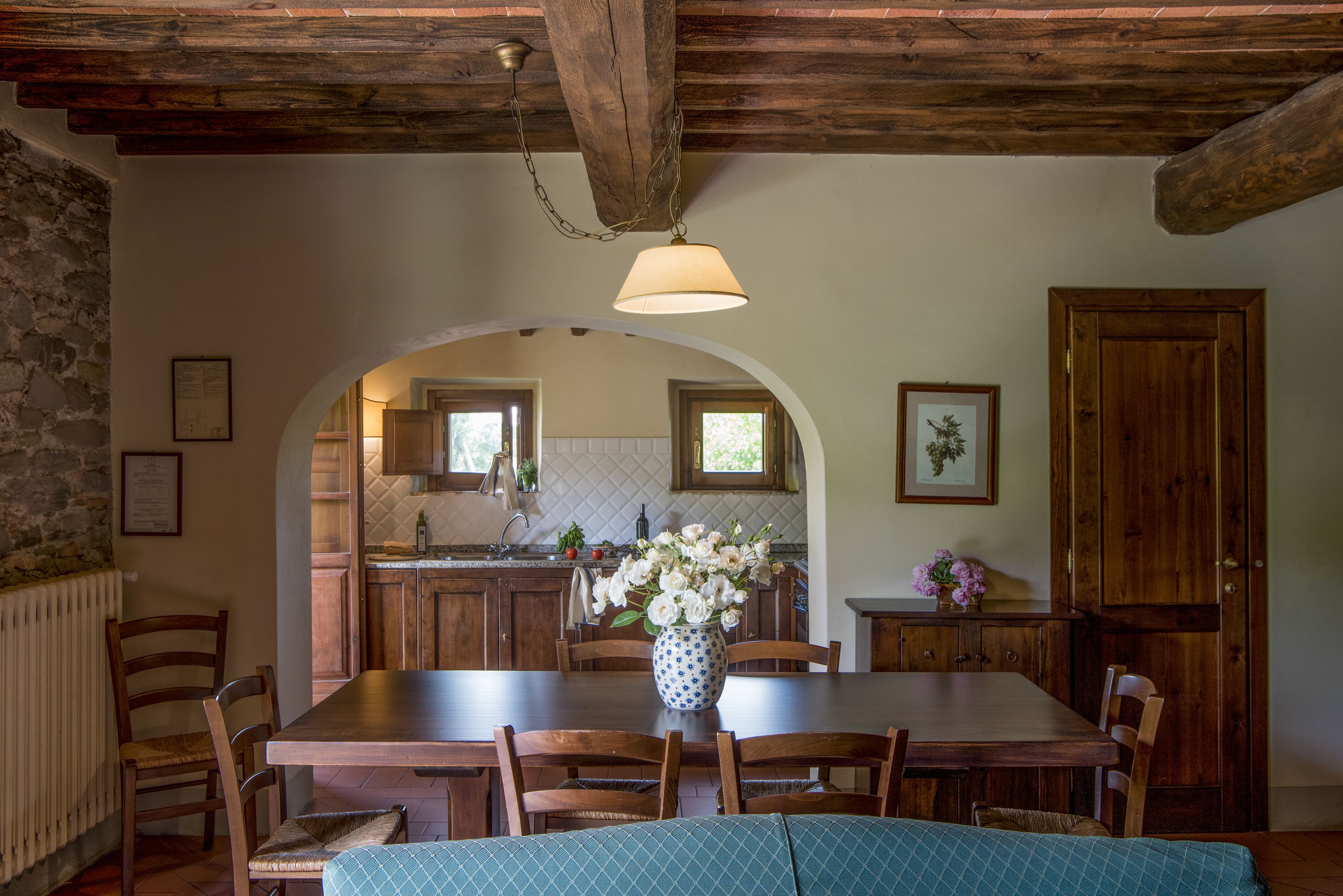 Un coin repas rustique avec une table en bois, des chaises et un vase de fleurs. On aperçoit une cuisine à travers une arche en arrière-plan.