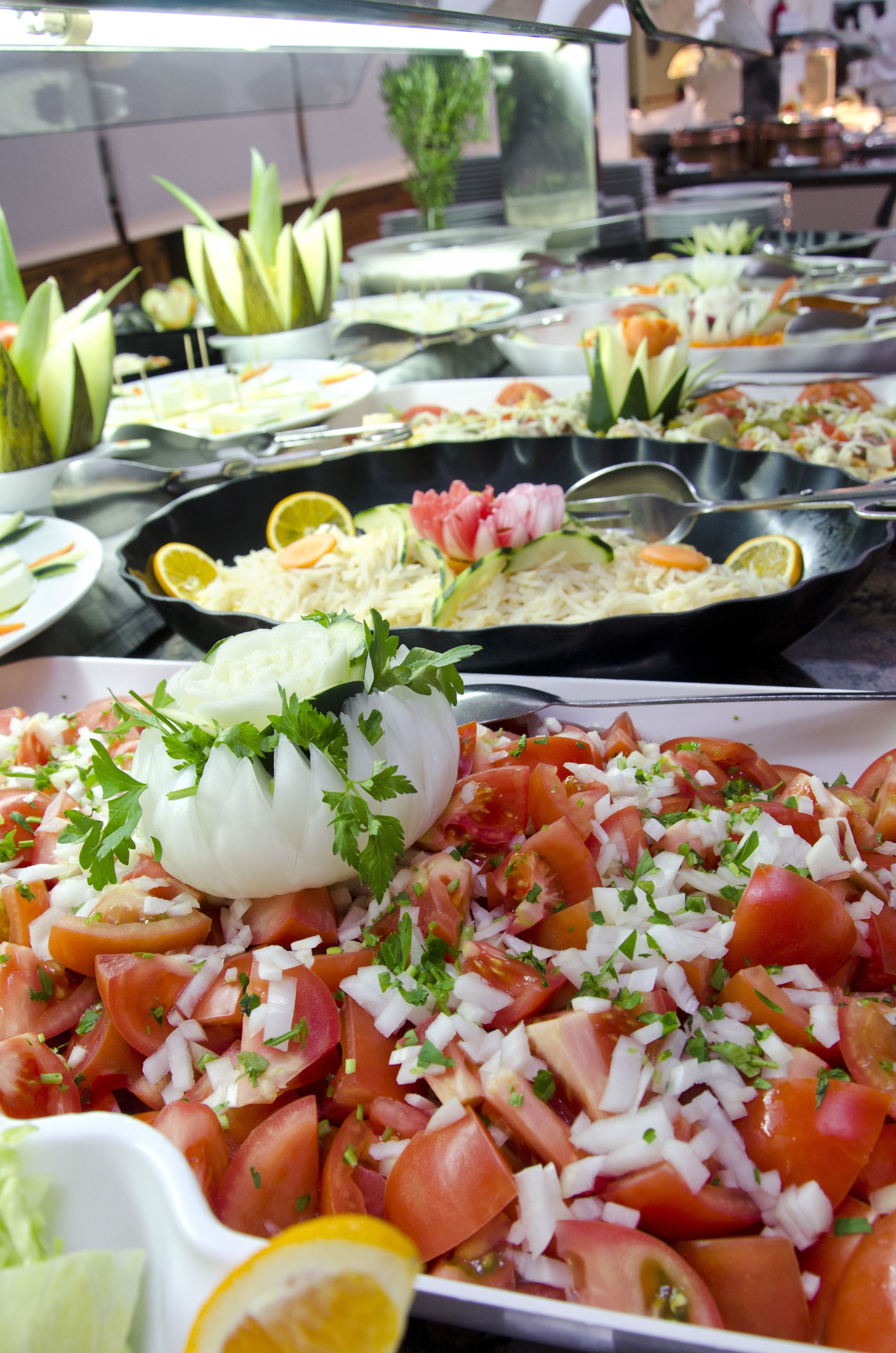Un bufé con diversos platos, incluyendo una ensalada con tomates, cebollas y hierbas, además de otros platos con verduras y guarniciones.