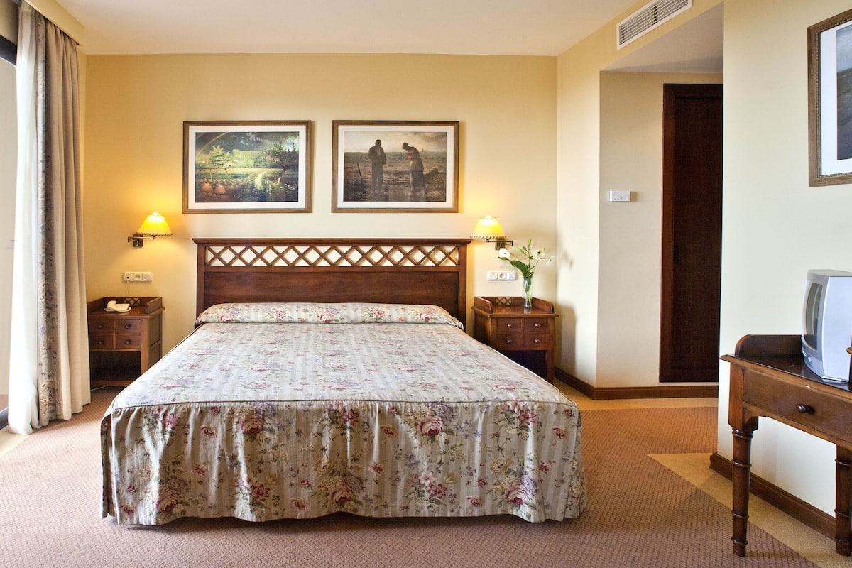 Una habitación de hotel con una cama grande, dos lámparas de pared, una mesita de noche de madera, una pequeña mesa con un televisor y una ventana con cortinas.