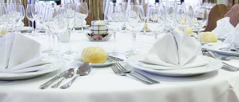 Una mesa de comedor dispuesta con manteles blancos, cubiertos y cristalería, con vistas al exterior a través de una ventana.