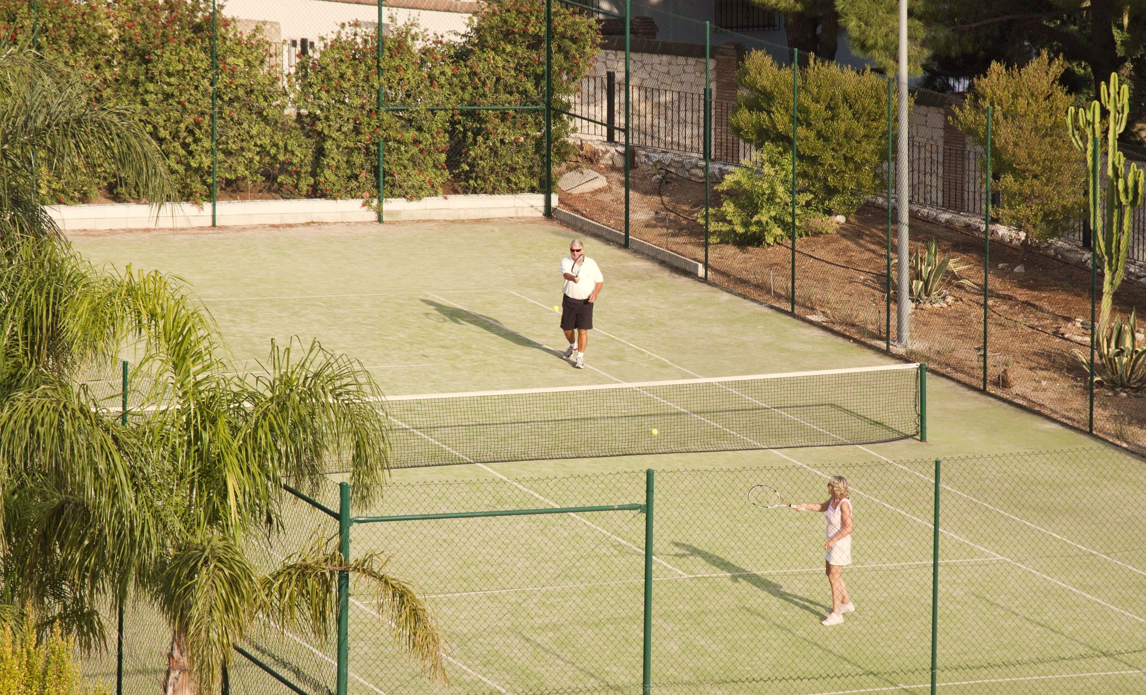 Una cancha de tenis con red, rodeada por una valla y árboles.