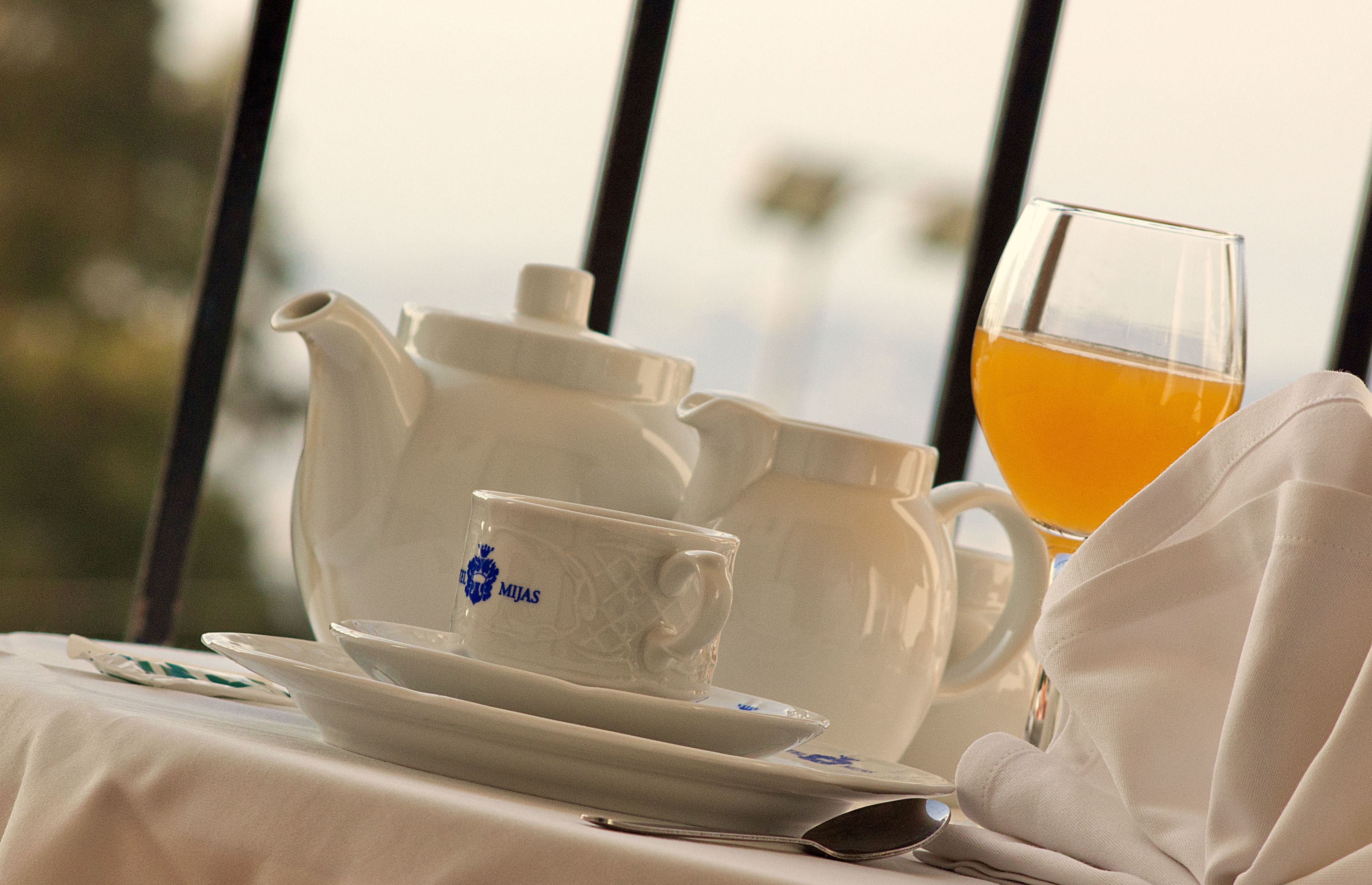 A tea set with a cup and saucer, a teapot, and a glass of tea on a table with a view through a window.