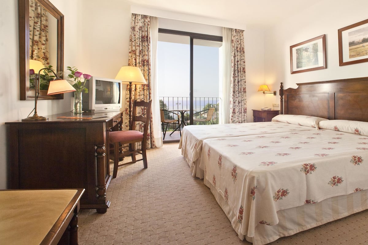 A hotel room with a large bed, a desk with a lamp, a chair, and a balcony with a view.