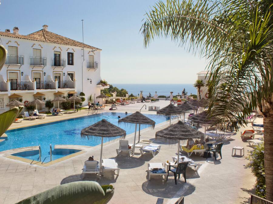 Zona de piscina con sillones de descanso y sombrillas, un edificio con balcones y vista al mar.