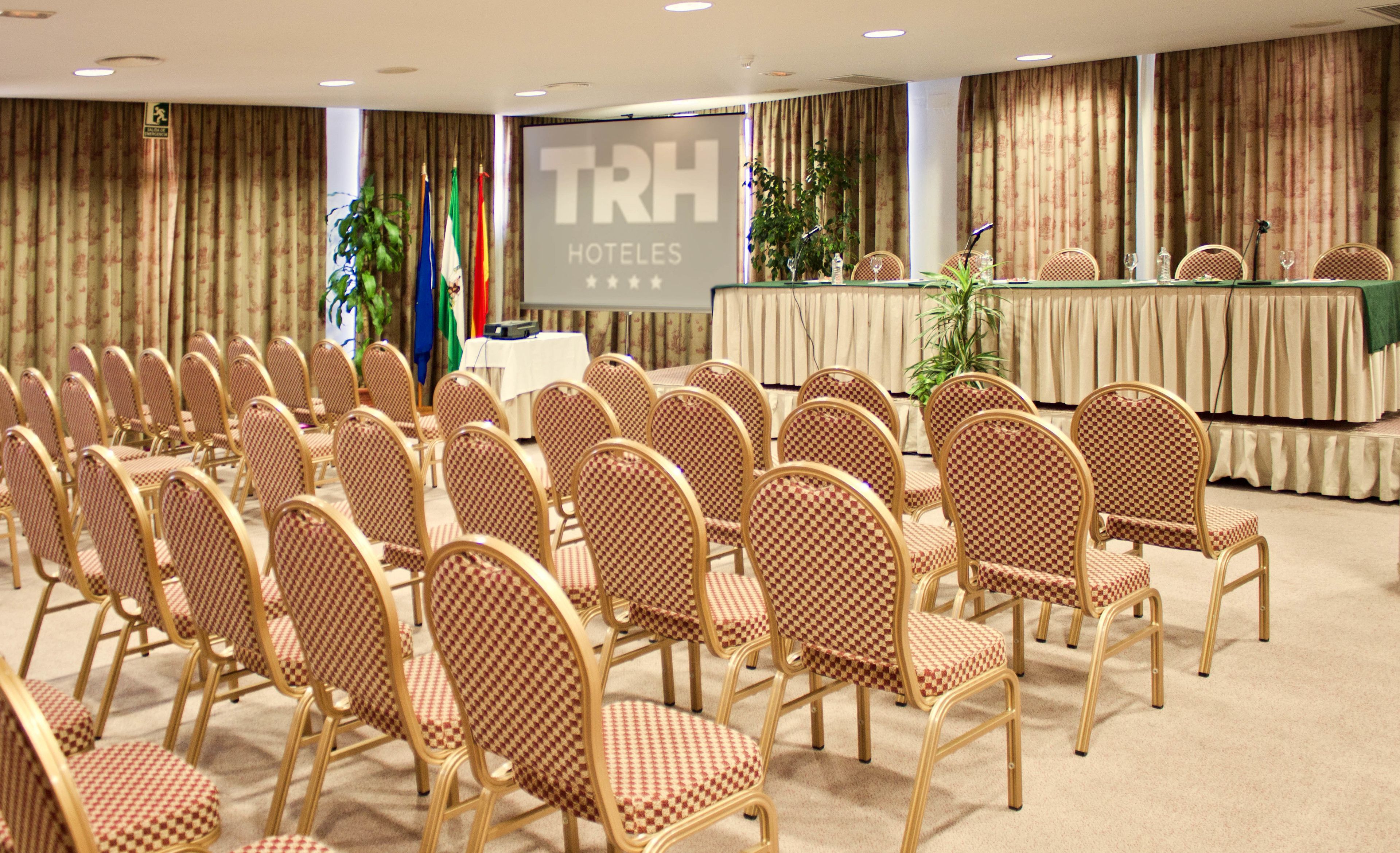Une salle de conférence avec des rangées de chaises à motifs à carreaux, une scène avec un pupitre et des drapeaux.