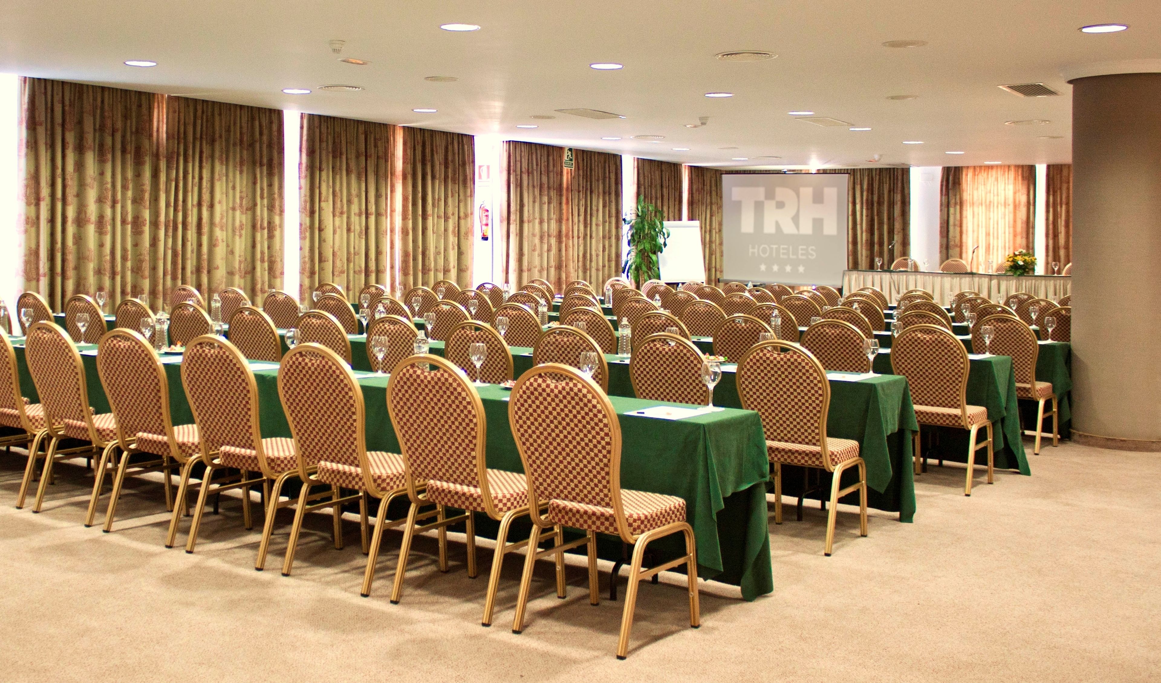 A conference room with rows of checkered chairs and tables, a projector screen, and a sign for TRH Hotels.