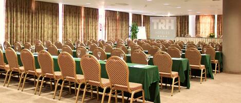 A conference room with rows of checkered chairs and tables, a projector screen, and a sign for TRH Hotels.
