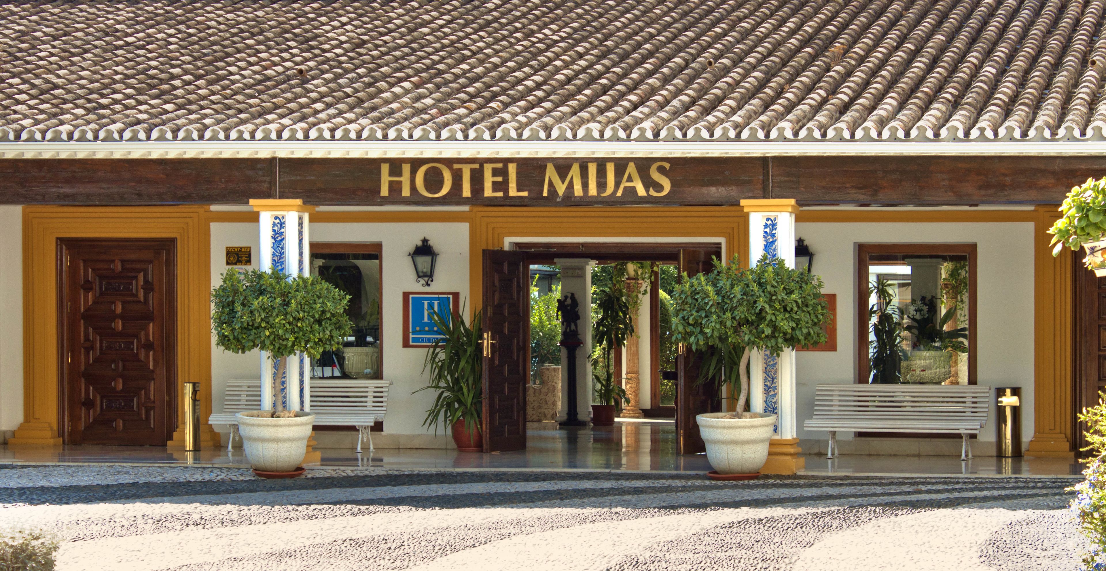 Entrada de un hotel con techo de tejas, un cartel que dice "HOTEL MIJAS" y plantas en macetas.