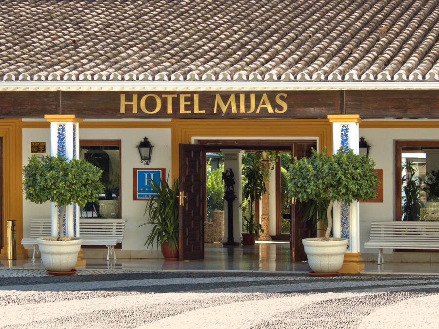Entrada de un hotel con techo de tejas, un cartel que dice "HOTEL MIJAS" y plantas en macetas.
