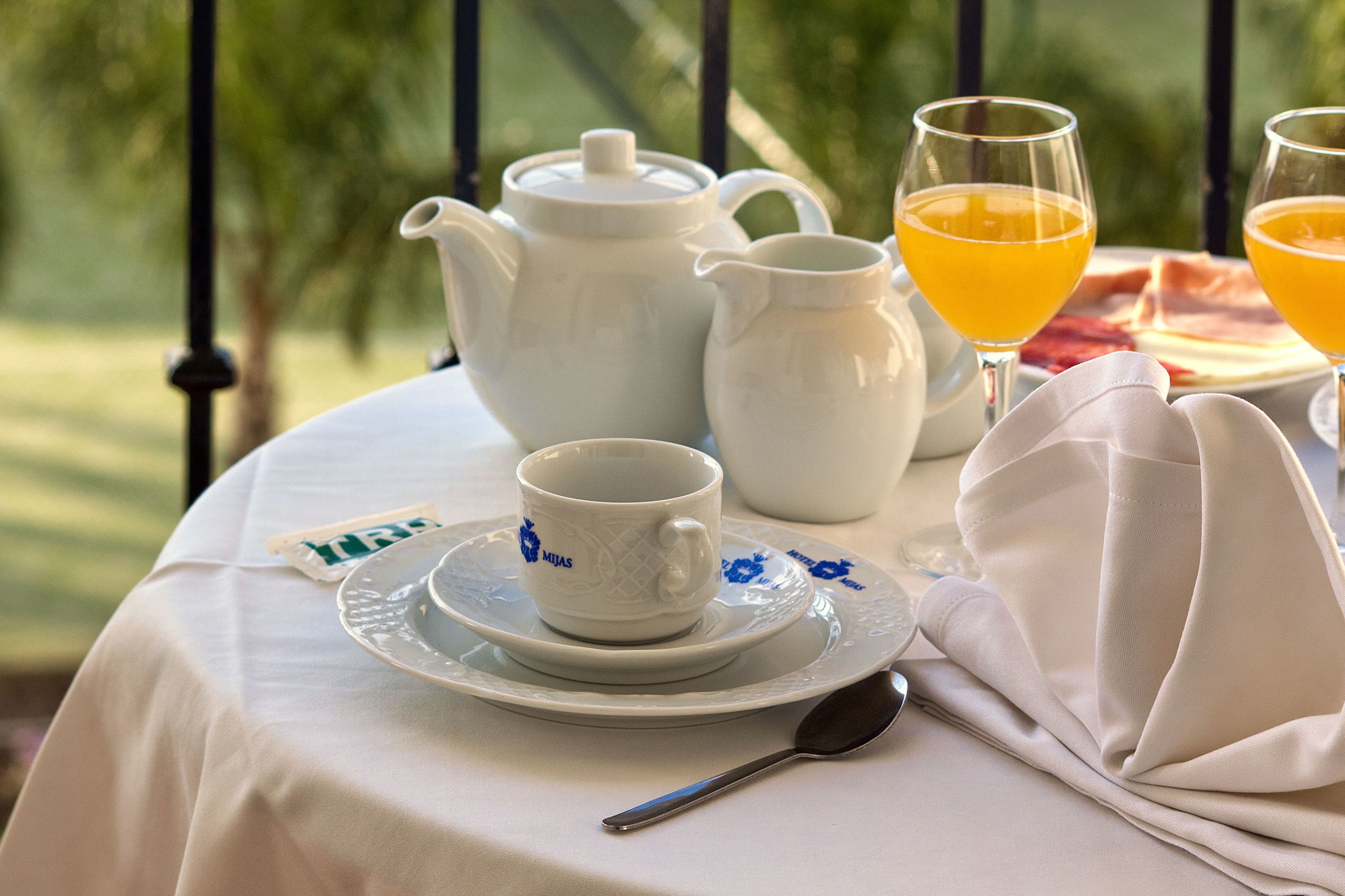 Ein Tisch ist gedeckt mit einem weißen Teekessel, Tassen und Gläsern Orangensaft, einem Löffel und einer Serviette, im Freien mit Blick auf Grünflächen.