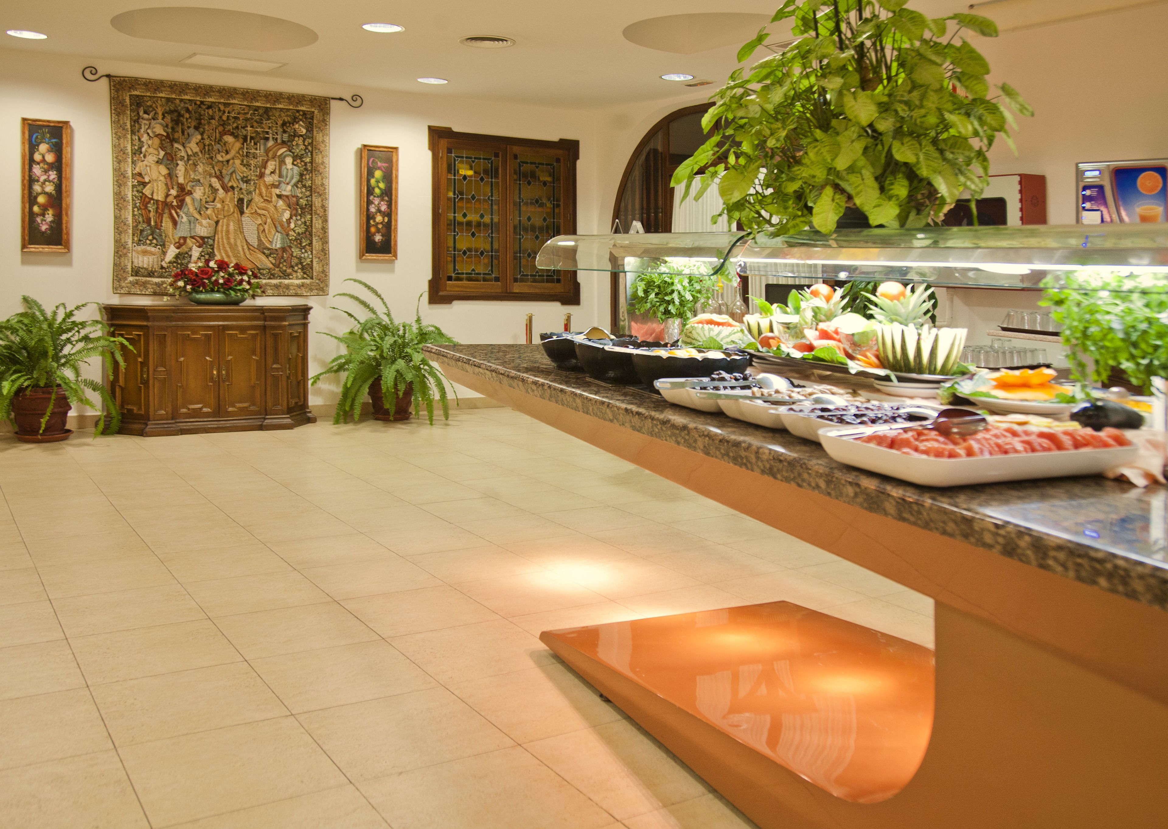 Un área de bufé con gran variedad de alimentos, una planta grande en maceta y un cuadro decorativo en la pared.