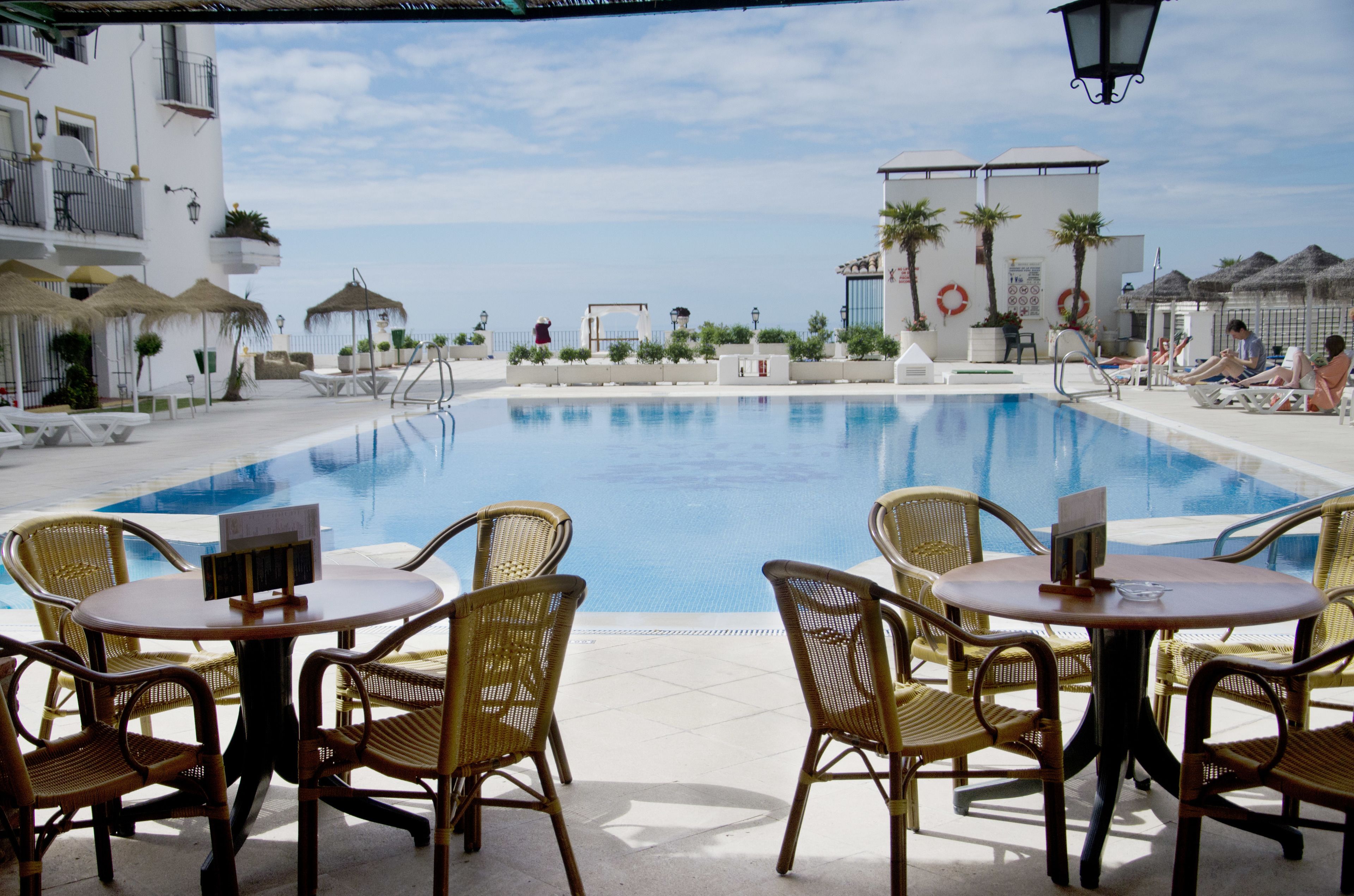 Zona junto a la piscina con sillas y mesas de mimbre, vista despejada del agua y un edificio con balcones y palmeras.