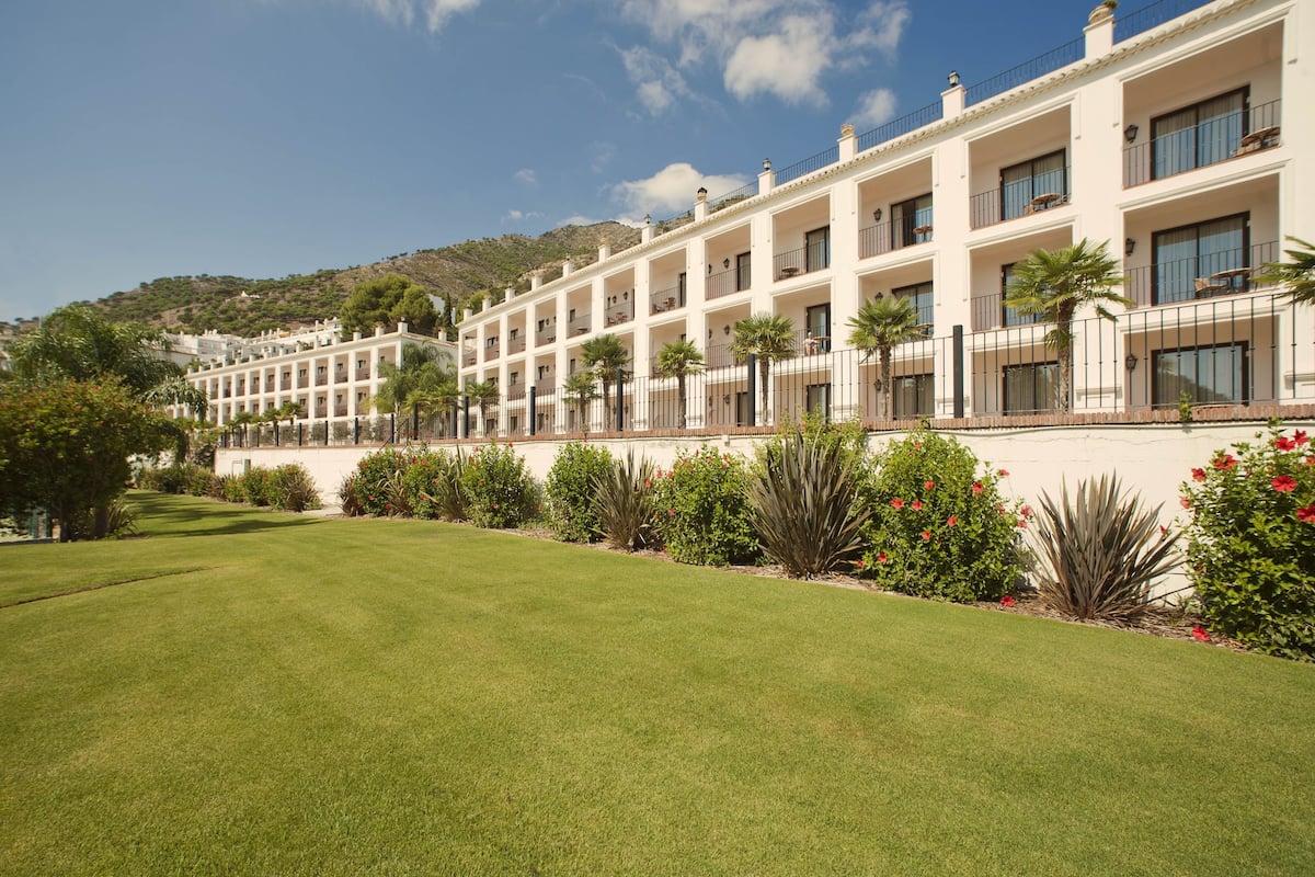 A multi-story hotel with a white facade, balconies, and a well-maintained lawn with various plants.