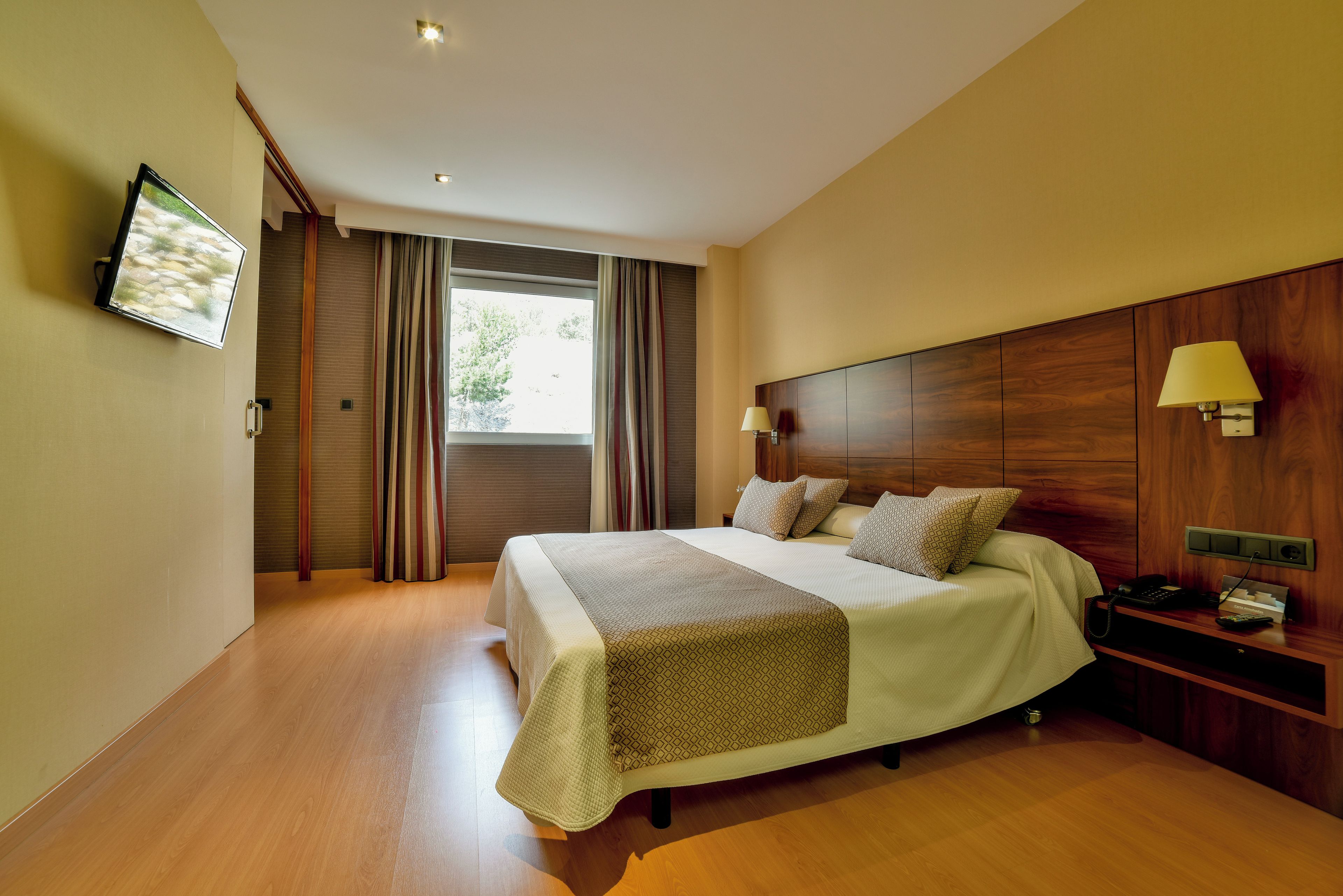 Una habitación de hotel moderna con una cama grande, un televisor montado en la pared y una mesita de noche con una lámpara y un teléfono.