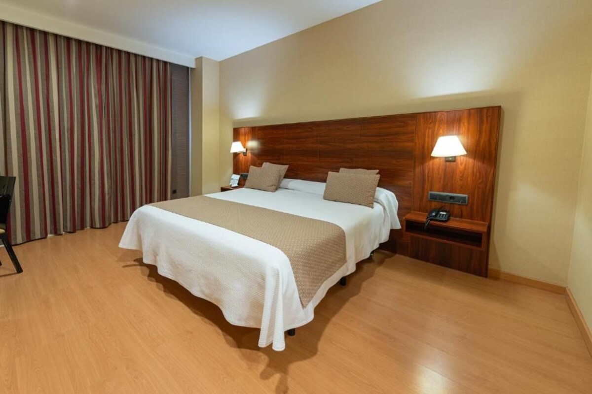 A hotel room with a large bed, wooden headboard, two bedside lamps, a telephone, and a curtain.