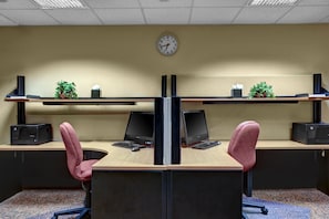 Two desks with computers, keyboards, and mice, and office chairs, set against a beige wall with floating shelves and potted plants.