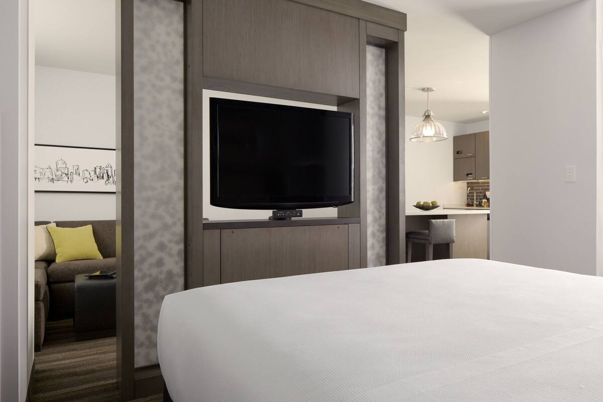 A modern hotel room with a large bed, a flat-screen TV mounted on a wooden panel, a small seating area with a sofa and a table, and a kitchenette in the background.