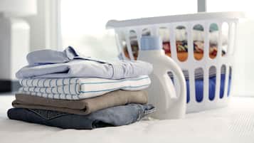 Una pila de ropa ordenada sobre una cama, con una cesta de ropa sucia al fondo.