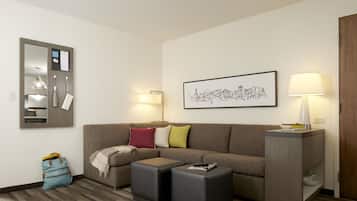 Ein modernes Wohnzimmer mit einer Eckcouch, zwei Hocker, einem Beistelltisch, einer Lampe, einem gerahmten Stadtbild und einem blauen Koffer auf dem Boden.