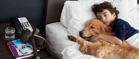 Una persona durmiendo en la cama con un perro, un control remoto, un vaso de agua y un libro en la mesita de noche.