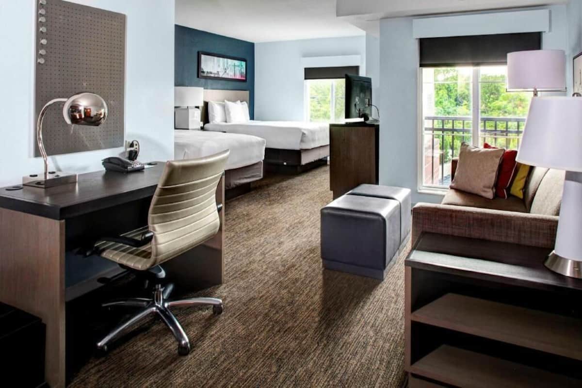 A hotel room with a desk, chair, two beds, a sofa, a TV, and a window with a view.
