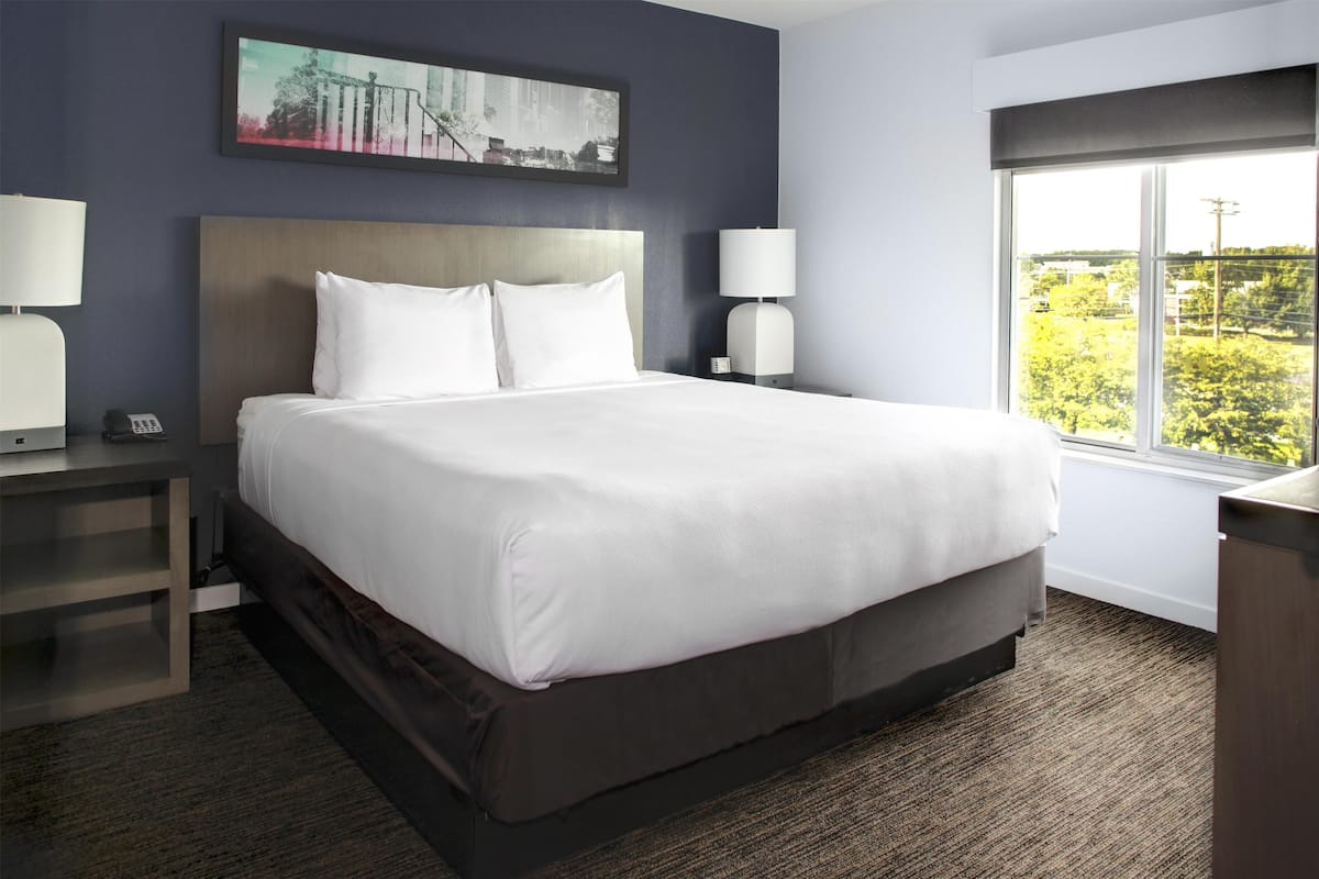 A hotel room with a large bed, two bedside lamps, a nightstand, a desk, and a window with a city view.
