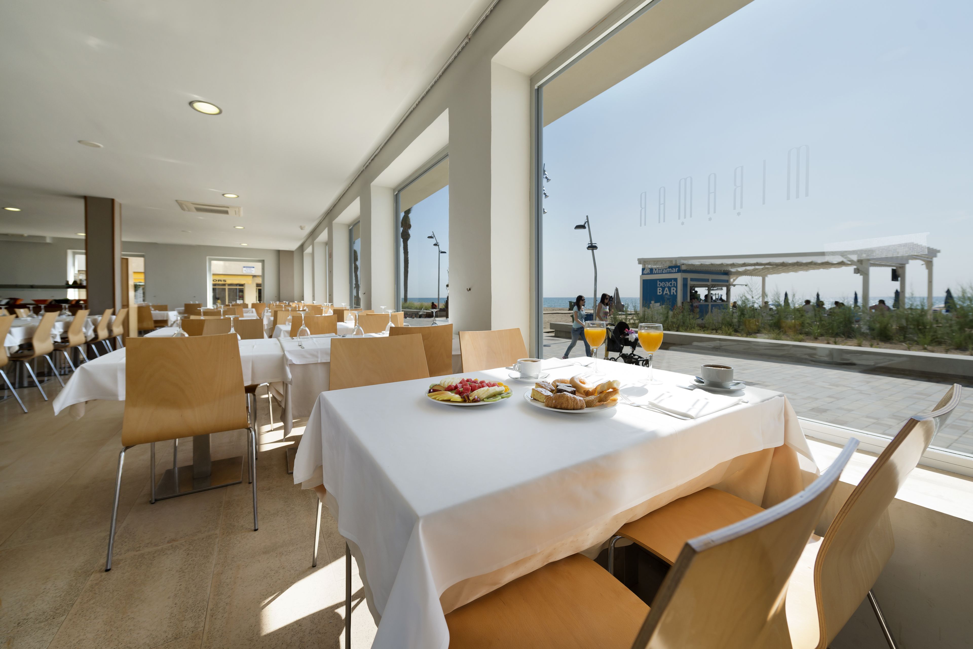 Amplia zona de comedor con mesas dispuestas para el desayuno, con vistas a una playa.