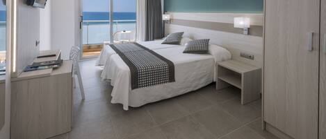 Habitación de hotel moderna con una cama grande, mesitas de noche, un escritorio con silla y vista al mar a través de una puerta corrediza de vidrio.