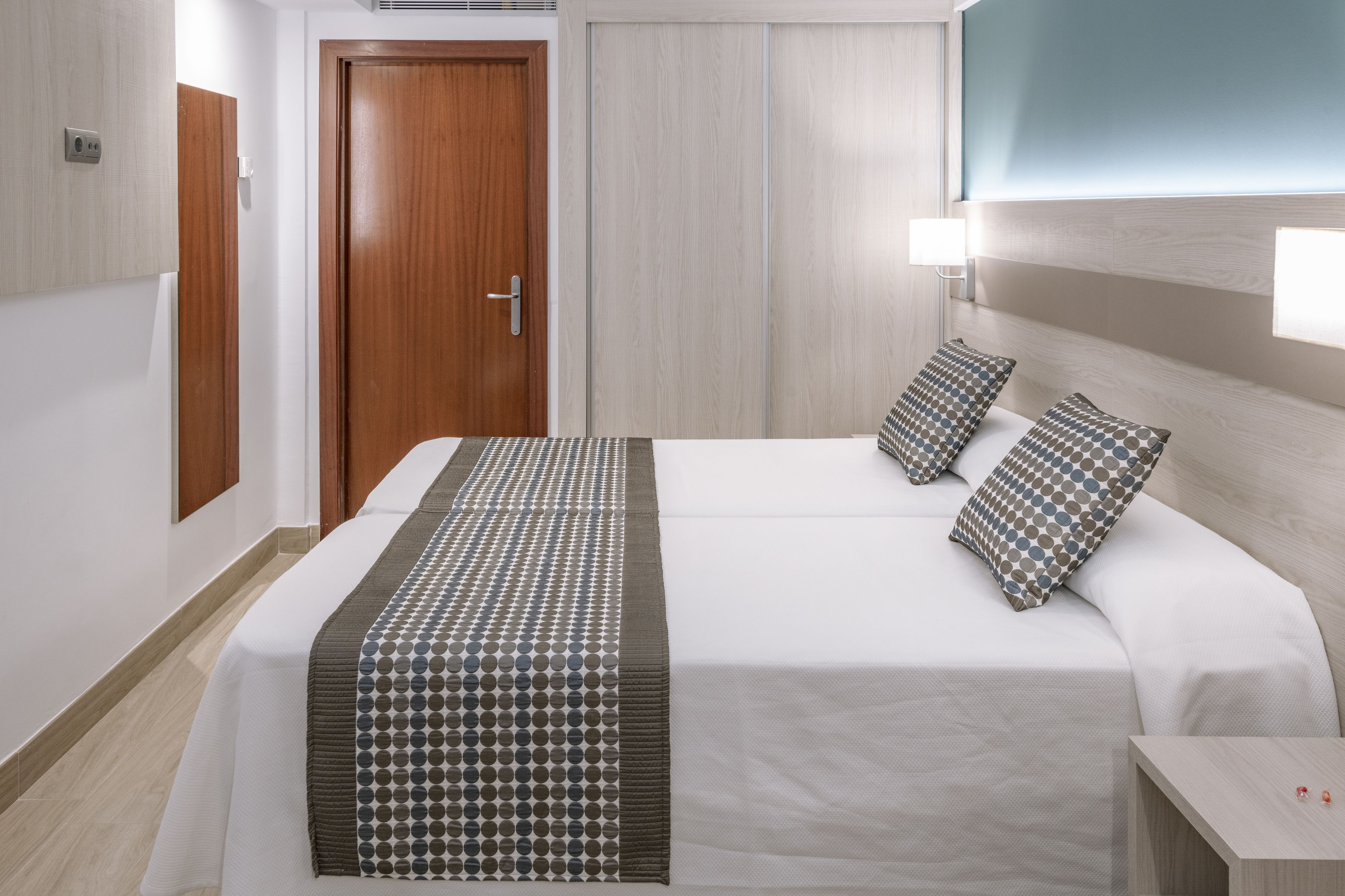 Una habitación de hotel con cama, dos almohadas, una mesita de noche y una lámpara de pared.