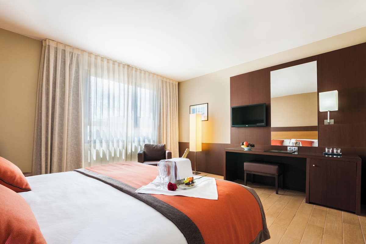 Ein Hotelzimmer mit einem großen Bett, einem Schreibtisch, einem Stuhl, einem Fernseher und einem Fenster mit Vorhängen.