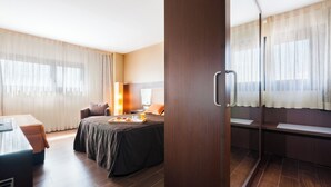A hotel room with a bed, a desk with a chair, a small table with breakfast items, and a window with curtains.