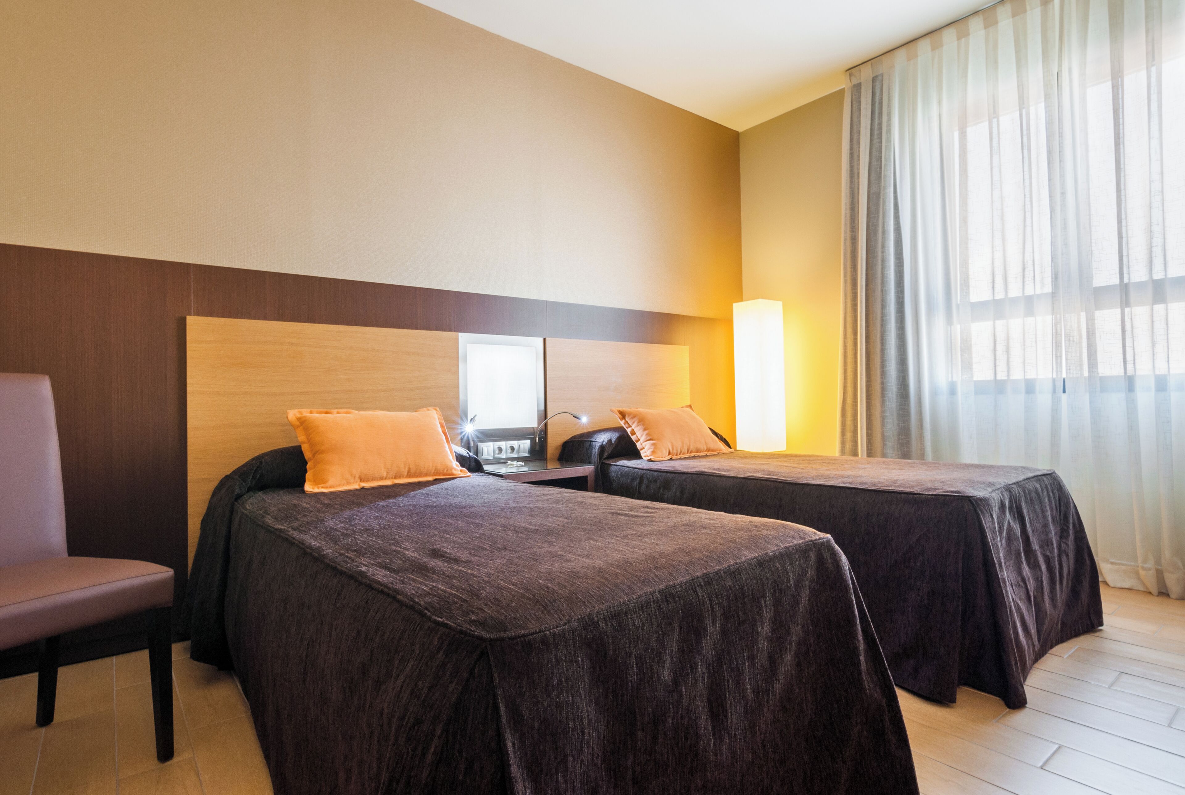 A hotel room with two beds, a wooden headboard, a chair, and a large window with curtains.
