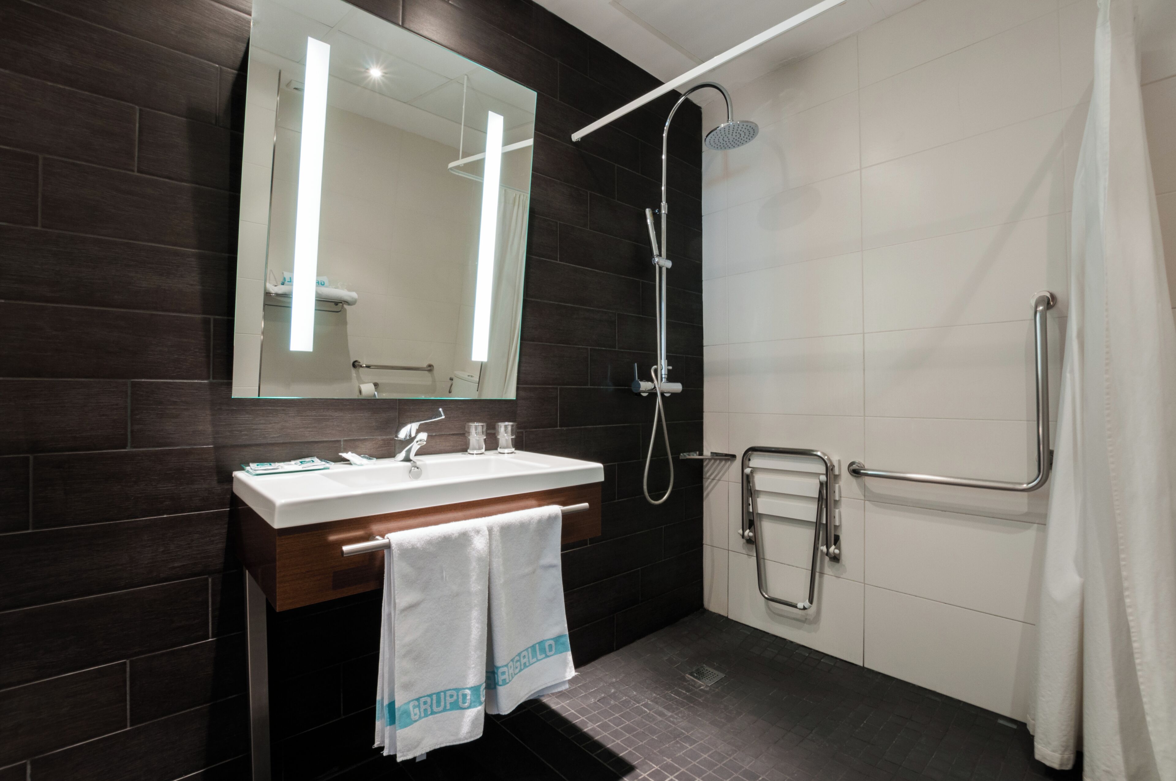 A modern bathroom with a dark wood vanity, a large mirror, a shower with a handheld showerhead, and a grab bar.